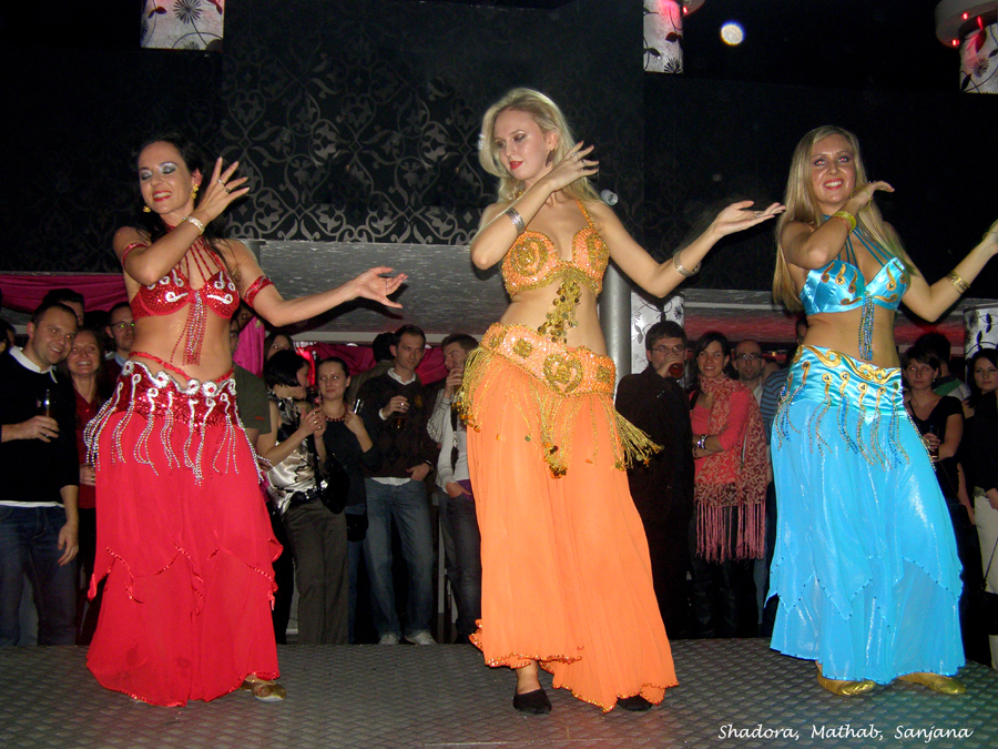 Trzy tancerki brzucha w barwnych, orientalnych strojach prezentują układ taneczny na scenie, otoczone publicznością w tle.